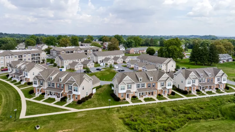 Aerial view of residential community featuring townhomes with varied siding, large green spaces, paved walkways, and tree-lined streets.