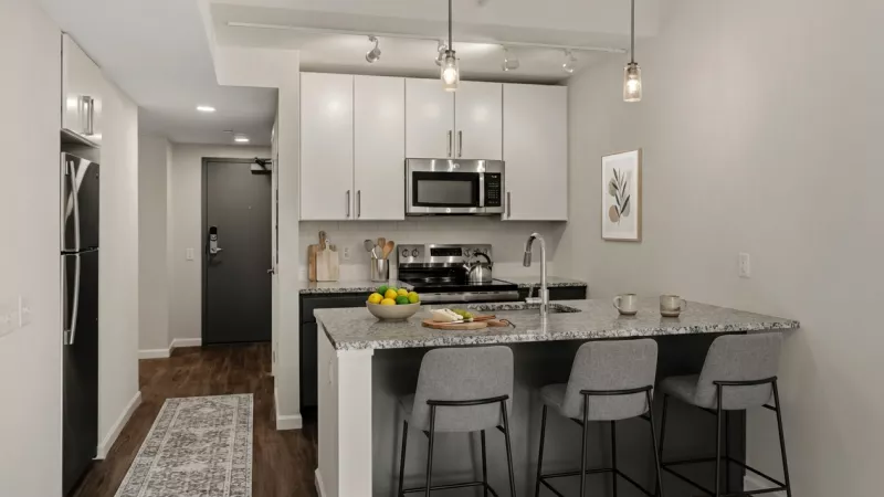 Sleek modern kitchen with light gray cabinets, stainless steel appliances, a speckled island with stools, and dark floor entryway.