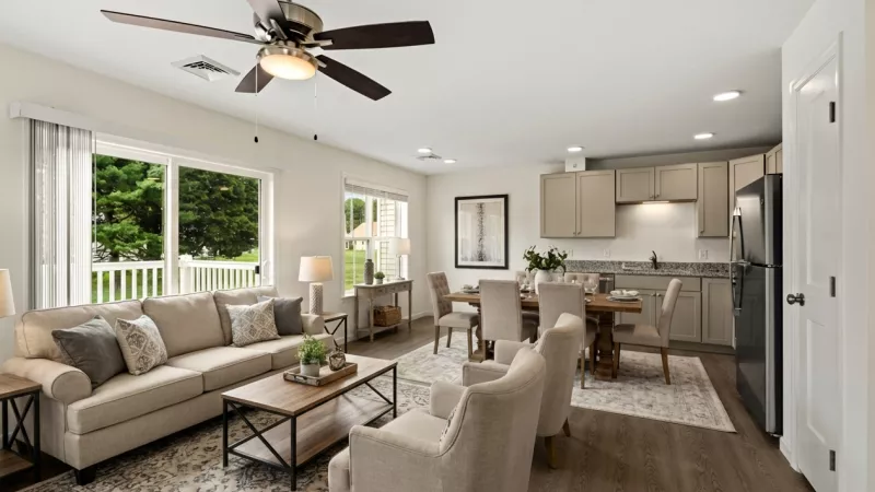 Bright open-plan living room with beige sofa, dining table, and kitchen featuring grey cabinets and stainless steel appliances.