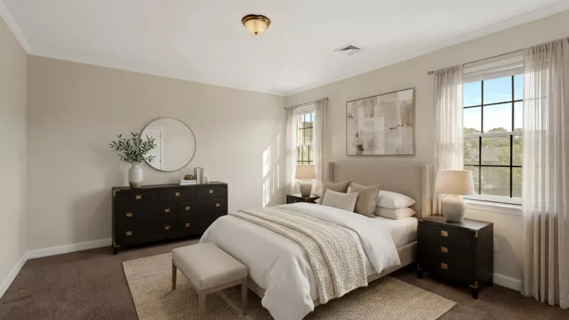 A bright, neutral-toned bedroom with a beige upholstered bed, dark wood dresser, matching nightstands, and two large windows.