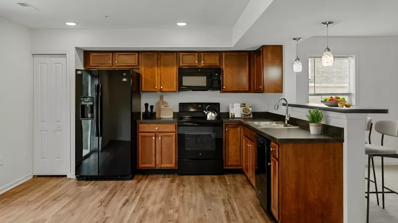 Modern kitchen with wood floors, brown cabinets, black appliances, dark counters, and a breakfast bar with stools.