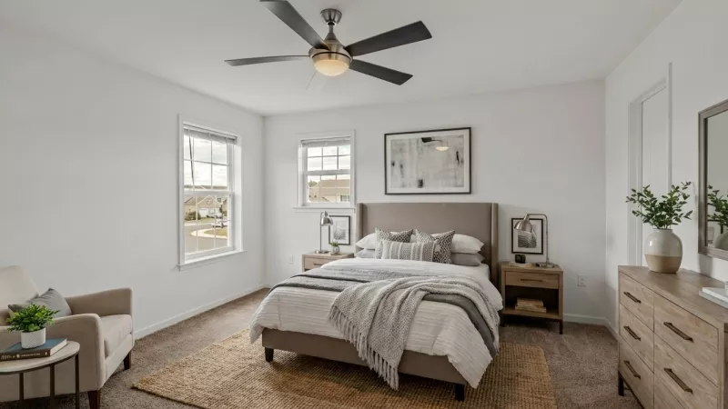 Bright, neutral-toned bedroom with a bed, grey headboard, white bedding, nightstands, dresser, armchair, and two windows.