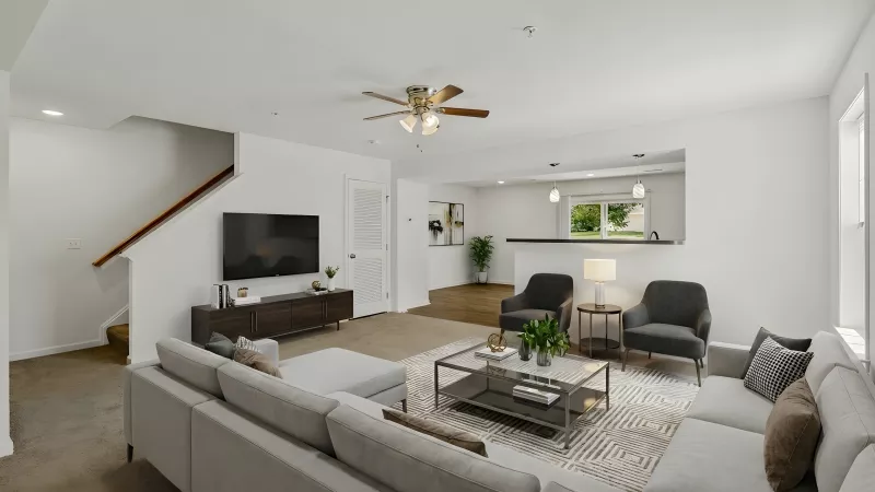 Bright, open living room with gray sectional, coffee table, TV, and staircase, looking into a kitchen.