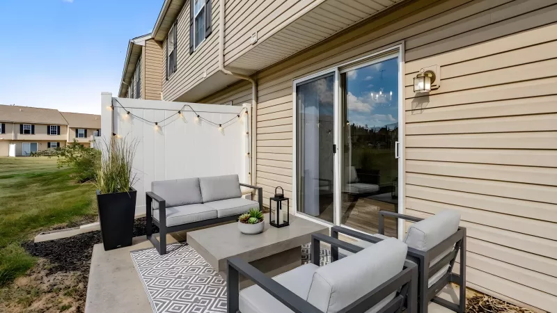 Outdoor patio with a gray sofa, two chairs, concrete table, string lights, and a sliding glass door next to light brown siding.