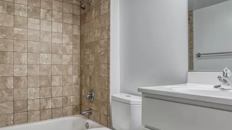Bathroom with white tub, toilet, vanity, beige square tiles above tub, and wood-look floor.