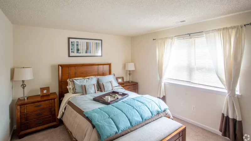 Furnished bedroom with a wooden bed, light blue and brown bedding, two nightstands with lamps, and a window with drapes.