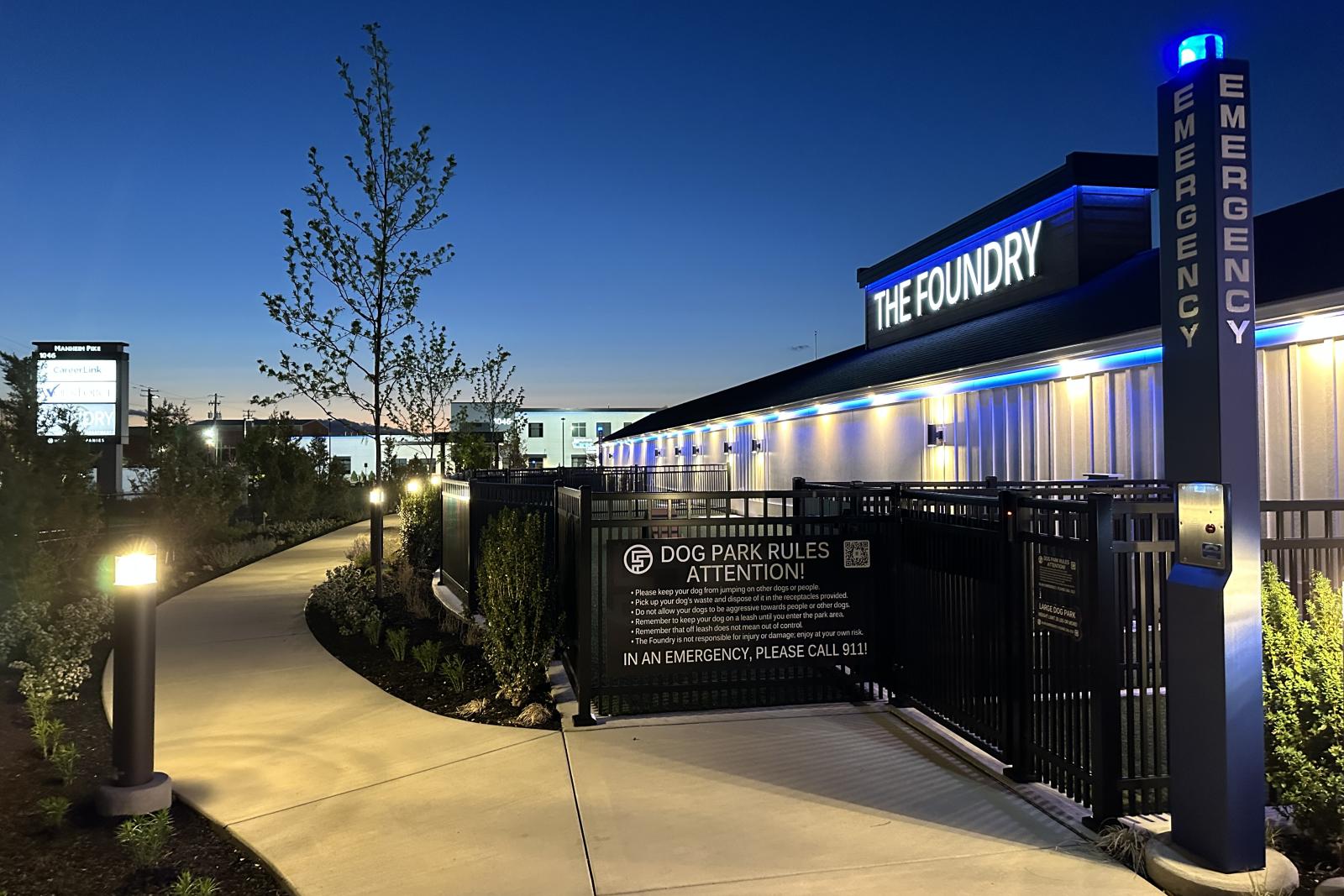 Nighttime view of The Foundry’s fenced dog park entrance, lit pathway, and signage with emergency contact information and pet rules.