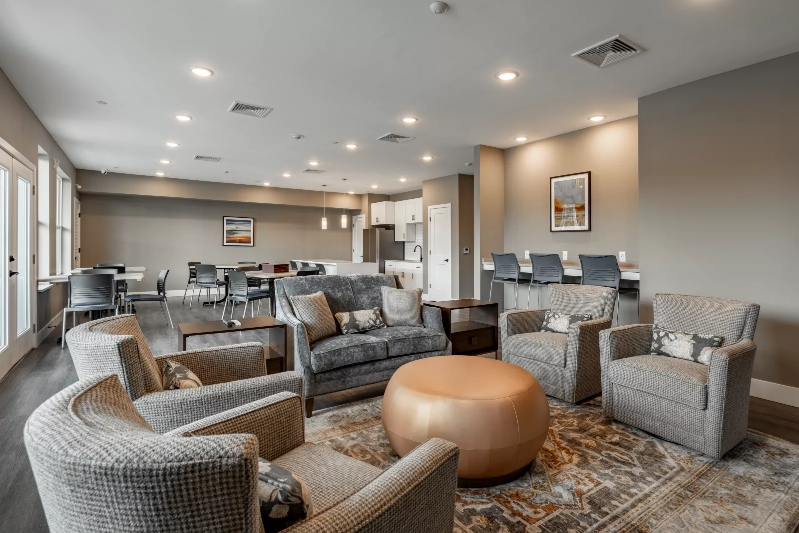 omfortable lounge seating area with plush gray armchairs, a velvet sofa, and a round ottoman, set on a patterned rug near a cozy community kitchen