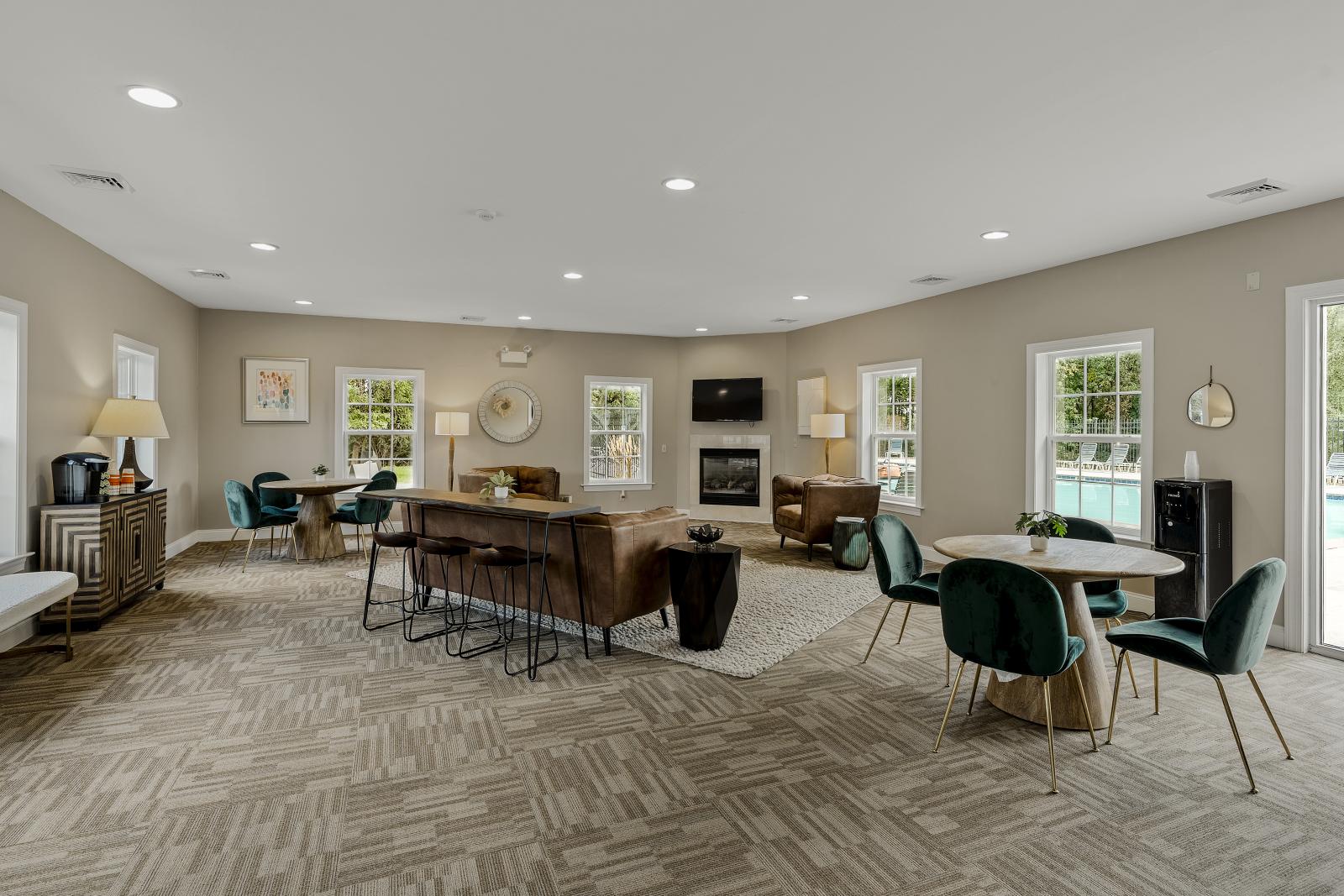 The interior of the resident clubhouse at Keystone Arms in Carlisle, PA. The photo features a lounge area with comfortable leather couches, coffee tables, and a cozy fireplace with a mounted television. There are also several small tables and chairs for working or socializing, with large windows providing a view of the pool deck. This image showcases the inviting community space available for residents.