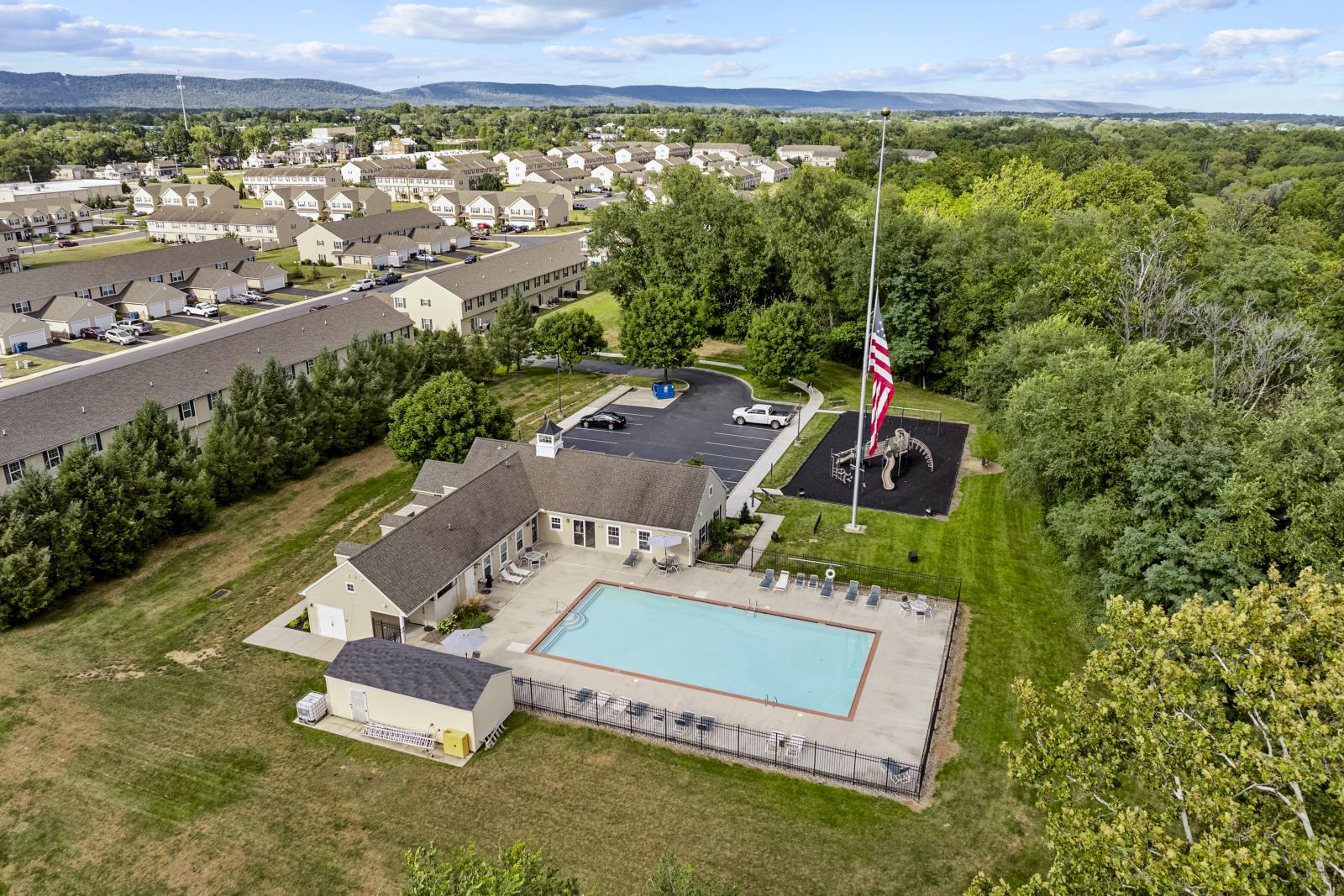 An aerial drone view of the clubhouse and swimming pool at Keystone Arms in Carlisle, PA. The photo shows the resort-style pool with a large sun deck, lounge chairs, and the community clubhouse building. In the background, there is a parking lot and a playground, all set against a backdrop of lush green trees.
