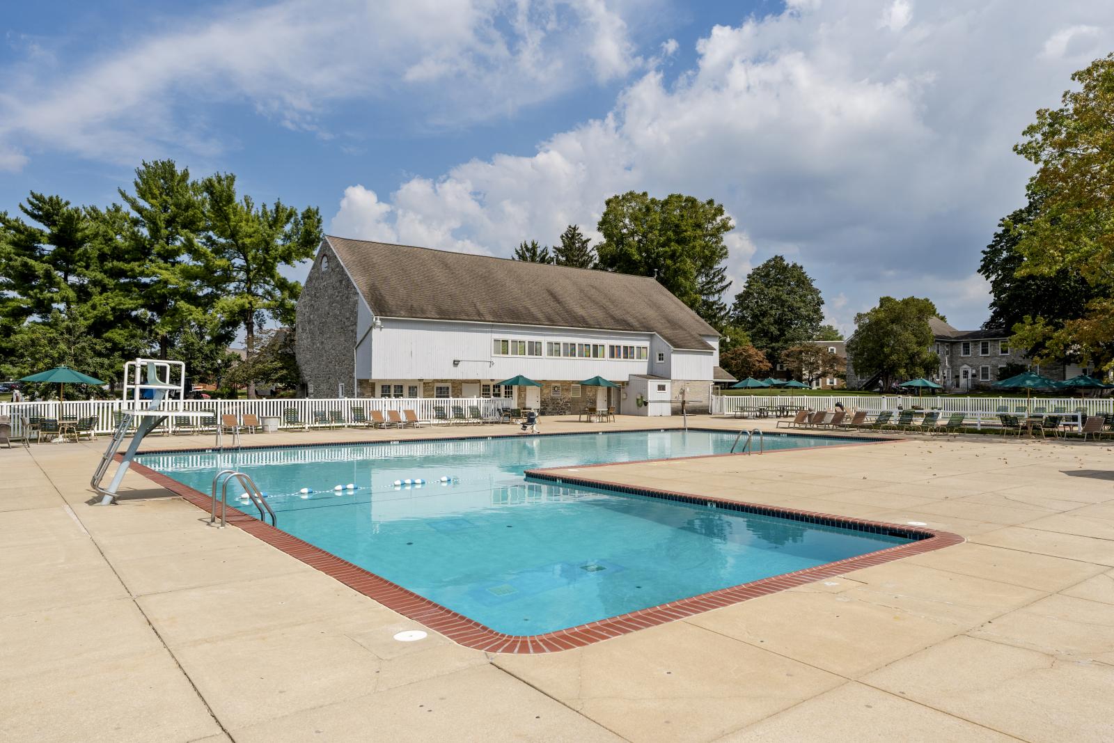 Large outdoor swimming pool with lounge chairs and umbrellas, set beside a barn-style clubhouse surrounded by trees and blue skies.
