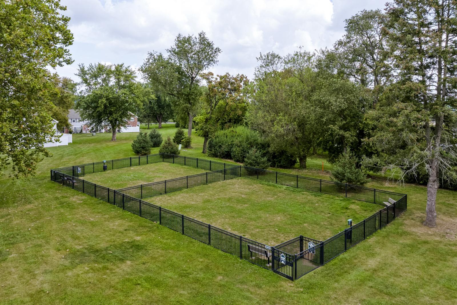 Fenced-in grassy dog park with benches and waste stations, surrounded by mature trees and open green lawns.