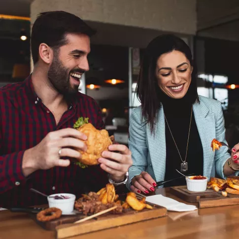 Smiling man and woman enjoying a casual meal together at a restaurant, featuring burgers, fries, and drinks, creating a fun and relaxed dining experience.