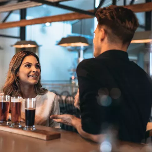 A bartender and a woman engaging in a friendly conversation at a stylish bar, with a flight of craft beers in front of her.