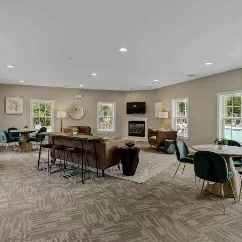 The interior of the resident clubhouse at Keystone Arms in Carlisle, PA. The photo features a lounge area with comfortable leather couches, coffee tables, and a cozy fireplace with a mounted television. There are also several small tables and chairs for working or socializing, with large windows providing a view of the pool deck. This image showcases the inviting community space available for residents.