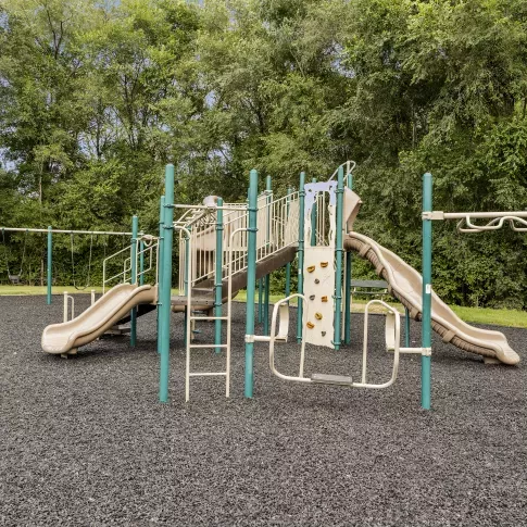 A view of the community playground at Keystone Arms in Carlisle, PA, featuring swings, slides, climbing equipment, and a large, mulched play area surrounded by mature trees. 