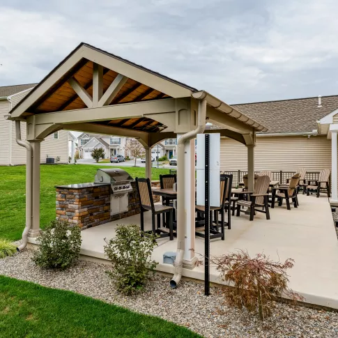 A wide-angle view of a concrete community patio featuring a covered grilling area and several rows of Adirondack-style lounge chairs. The space is situated behind residential buildings with light-colored siding and overlooks a shared green space.