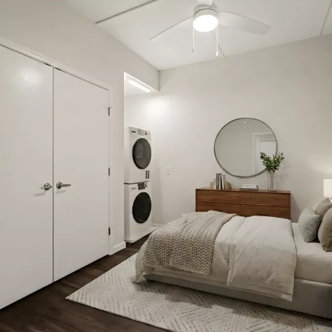 Modern bedroom with a bed, wood dresser, round mirror, and a stacked washer/dryer. Dark wood floor, light walls, and ceiling fan.