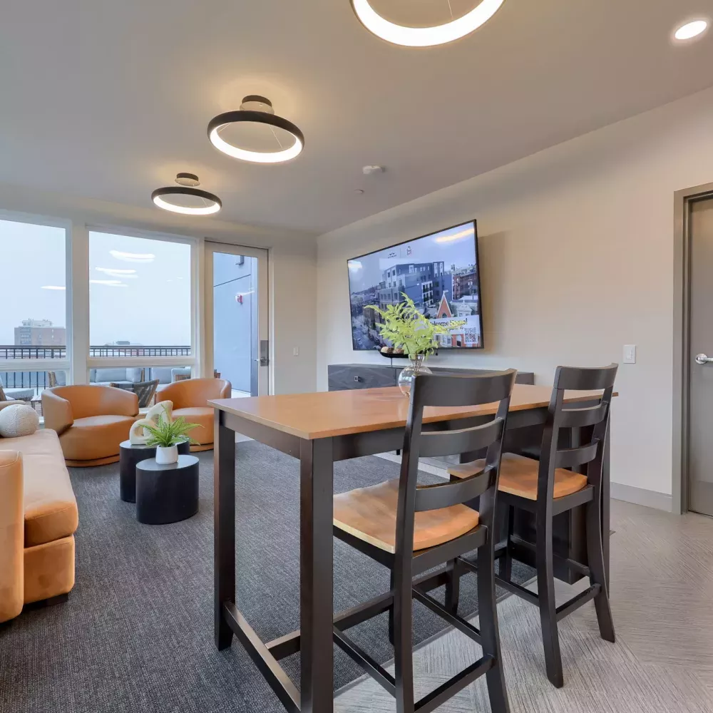 Bright, modern lounge with orange sofas, a wooden high-top table with chairs, large windows, and a wall-mounted TV.