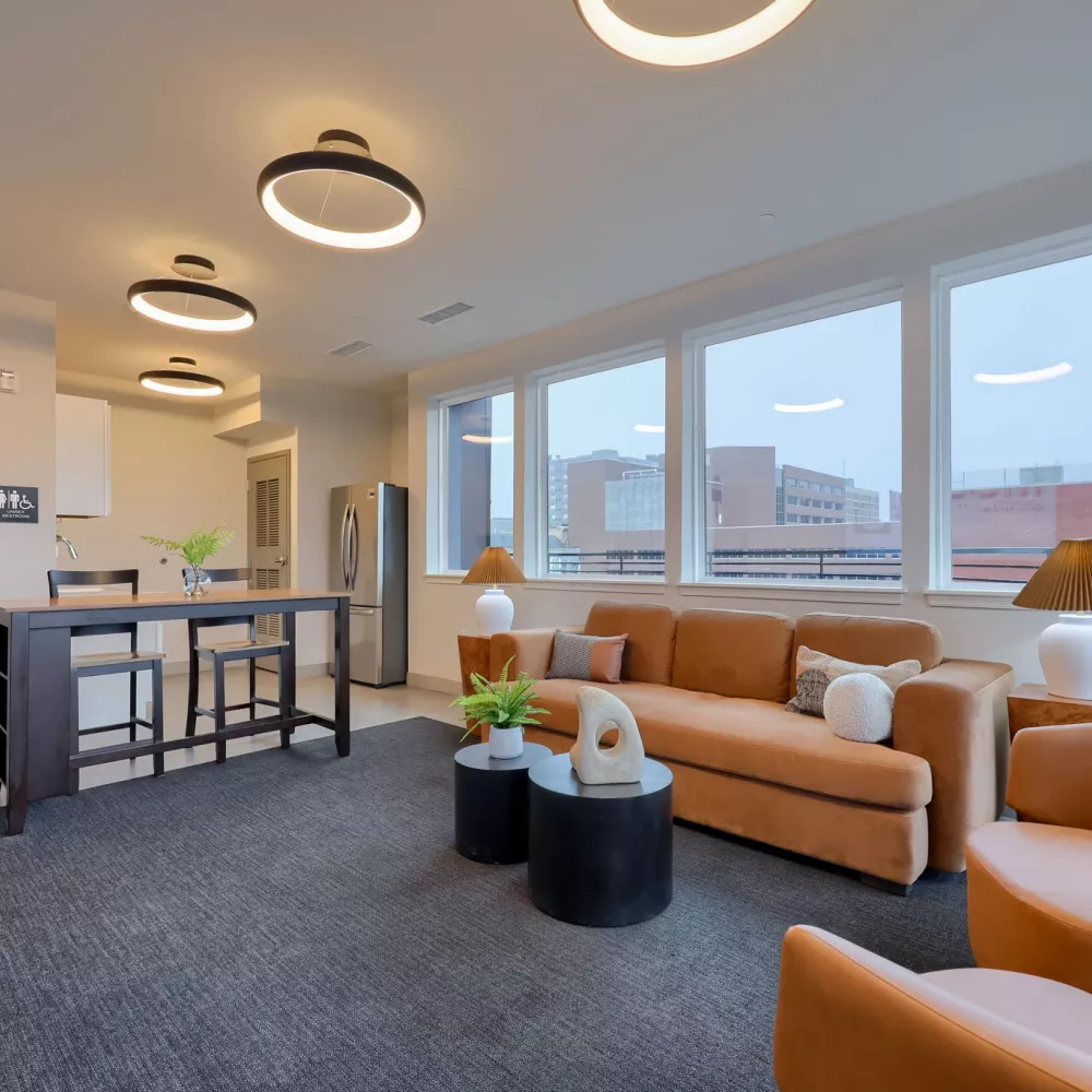 Modern open-plan living room and kitchen with orange furniture, large windows, dark gray rug, and circular ceiling lights.