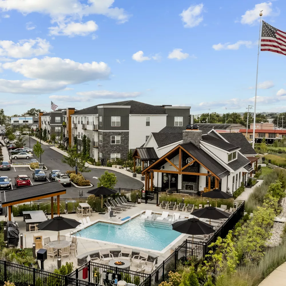 Aerial photograph of a modern apartment complex featuring a swimming pool, clubhouse, and an American flag against a blue sky.