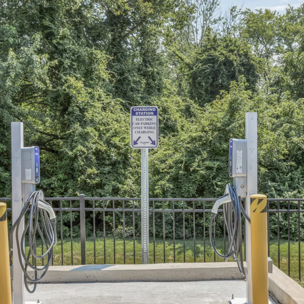 Outdoor EV charging station with two chargers, a sign, yellow bollards, a black fence, and green trees in the background.