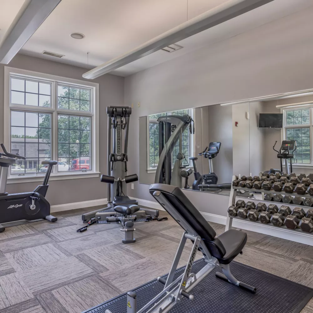 Photograph of a bright fitness center with exercise bikes, a weight bench, and a large rack of dumbbells, reflecting in a wall mirror.
