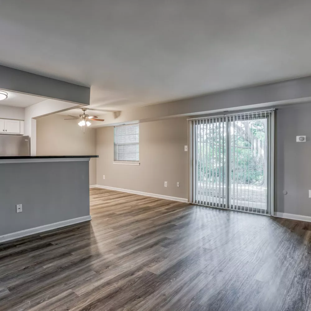Empty living room with wood floors, grey walls, and a pass-through to a kitchen. Sliding glass door leads outside.