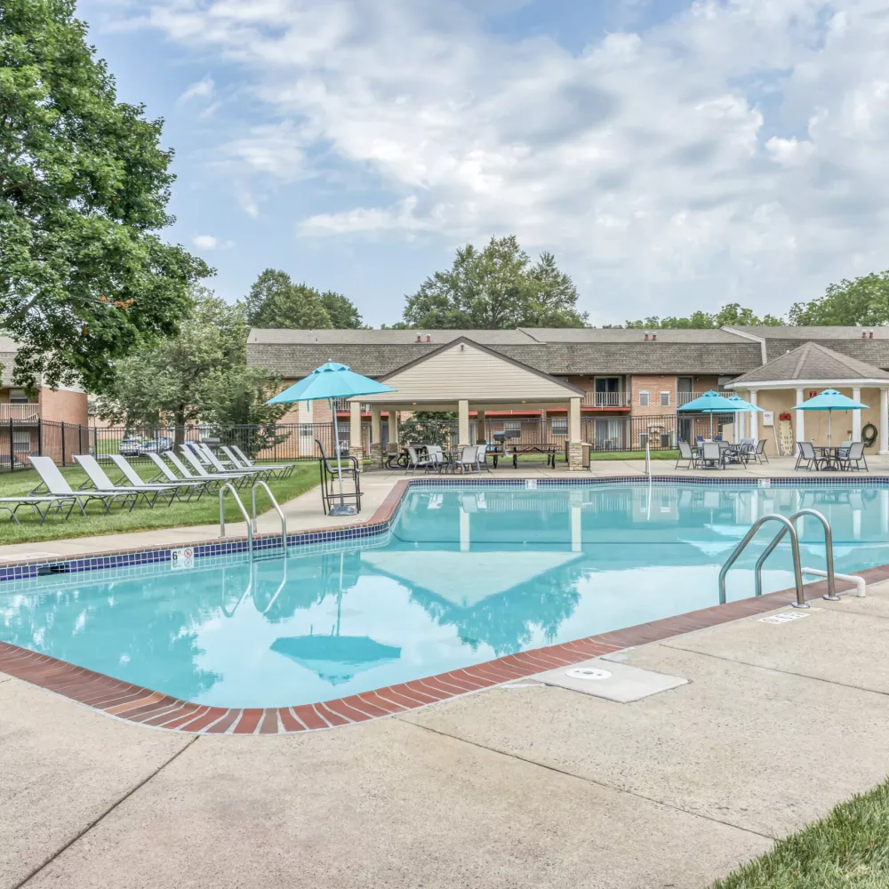 Outdoor swimming pool with lounge chairs, covered patio, blue umbrellas, and apartment buildings under a partly cloudy sky.
