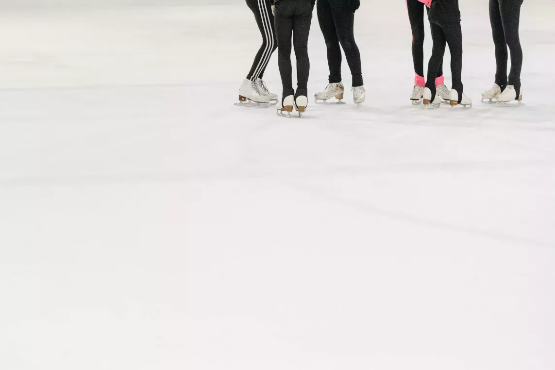 Group of figure skaters wearing black leggings and ice skates standing together on an indoor ice rink during practice.