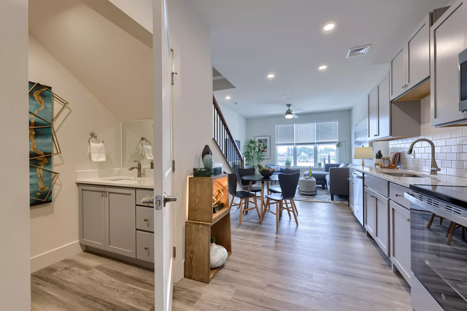 Open-concept kitchen and living area at The Pointe at Overlook with gray shaker cabinets, subway tile, stainless-steel appliances, round dining table, wood-style floors, and a powder room with quartz vanity off the hall.