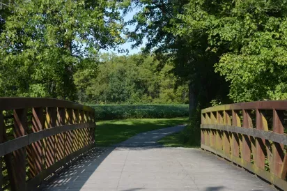 Paved walking over a Carlisle Creek with tall trees and open grassy areas in Carlisle, PA, on a sunny day.