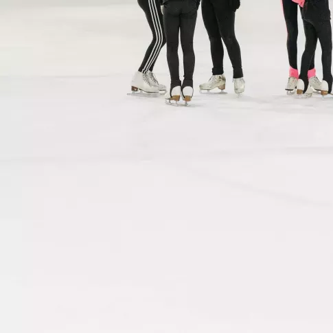 Group of figure skaters wearing black leggings and ice skates standing together on an indoor ice rink during practice.