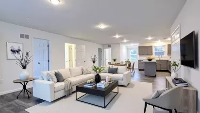A brightly lit, open-concept living space in a rental property. It features white walls, dark wood-look flooring, and a large white sectional sofa on a light-colored rug. The view extends into the kitchen with gray cabinetry and a dining area.