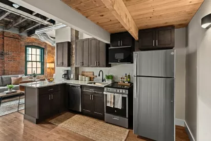 A modern kitchen with dark wood cabinetry, granite countertops, and stainless steel GE appliances. The design highlights industrial elements like exposed ductwork and rustic wooden ceiling beams.
