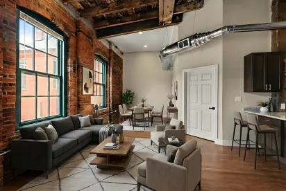 A spacious open-concept living area featuring original exposed red brick walls, towering industrial timber ceilings, and large arched windows that flood the room with natural light. The space includes a modern kitchen island with seating and a dining area.