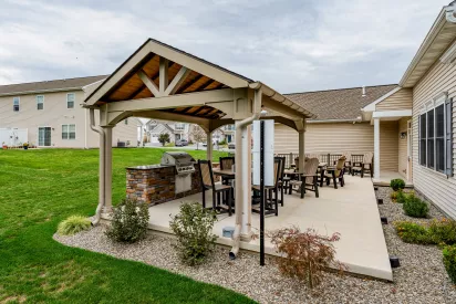 A wide-angle view of a concrete community patio featuring a covered grilling area and several rows of Adirondack-style lounge chairs. The space is situated behind residential buildings with light-colored siding and overlooks a shared green space.