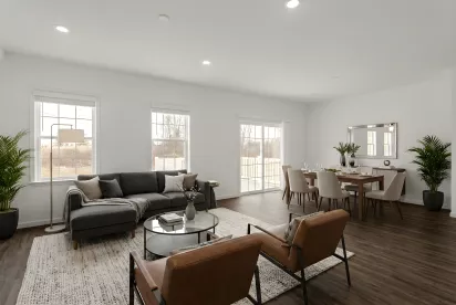 An expansive living and dining area with dark wood-style flooring and white walls. The room is illuminated by natural light from large windows and features a modern dining set and a comfortable seating area.