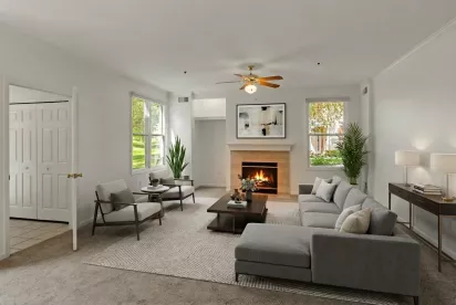 A spacious living room with neutral-toned carpeting, white walls, and a central fireplace with a tan stone surround and white mantel. The room includes a ceiling fan with integrated lighting and two large windows that provide a view of the exterior landscaping.