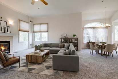 A spacious, open-concept living room and dining area featuring a fireplace, neutral carpeting, a large sectional sofa, and a dining table set for four near a large arched window.