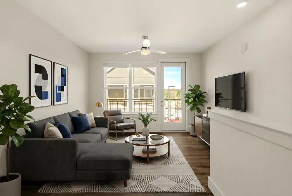 A bright living room with neutral walls and a large window and glass door providing natural light. The room is styled with a dark gray sectional, a round wooden coffee table, and a wall-mounted TV above a decorative ledge.