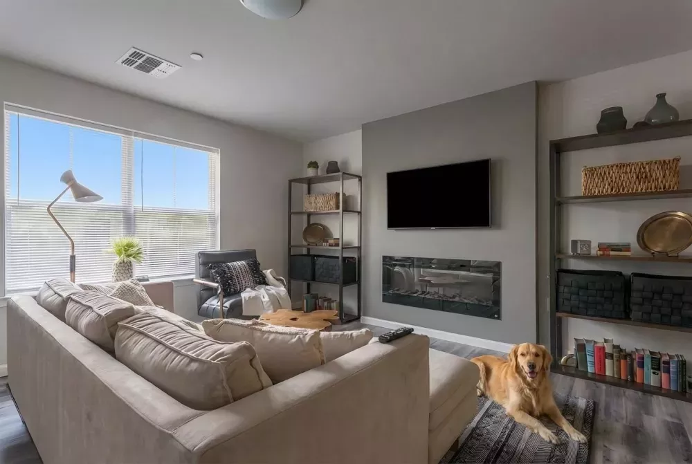 Modern living room with beige sectional sofa, TV above fireplace, industrial shelves, and a golden retriever on a patterned rug.