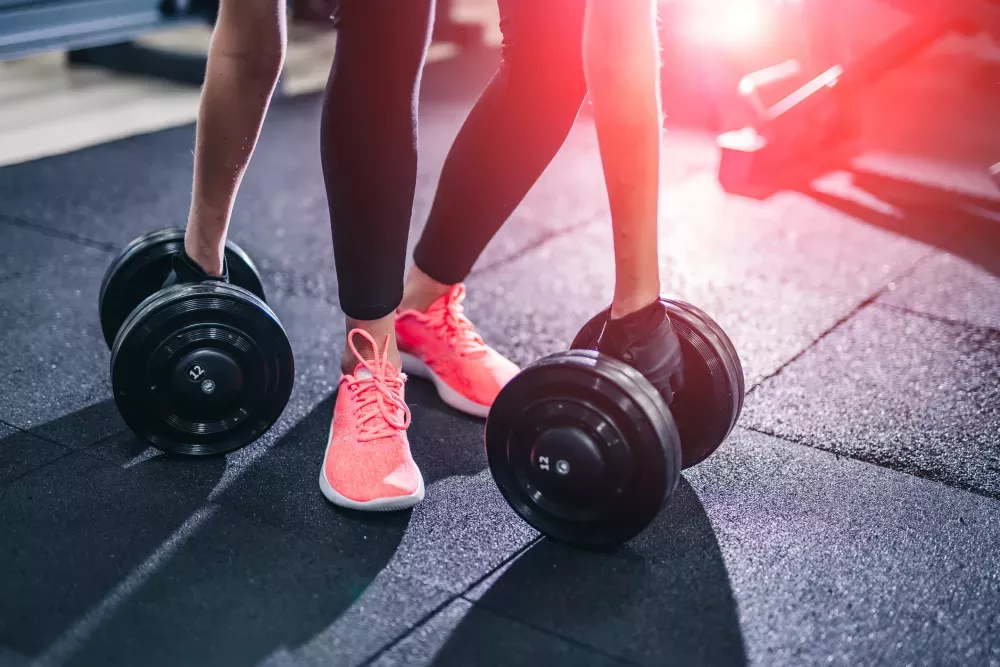 Person in pink sneakers reaches for a dumbbell on a dark gym floor, with another dumbbell nearby.