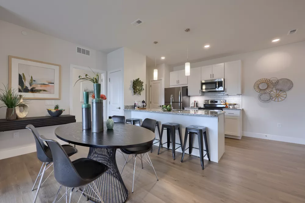 Modern open-concept apartment interior with a dining table, black chairs, and a kitchen with white cabinets.