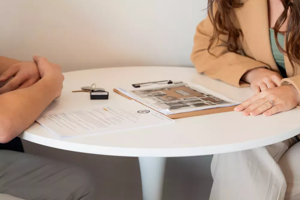 Two individuals at a table with real estate documents, keys, and a pen, discussing property.
