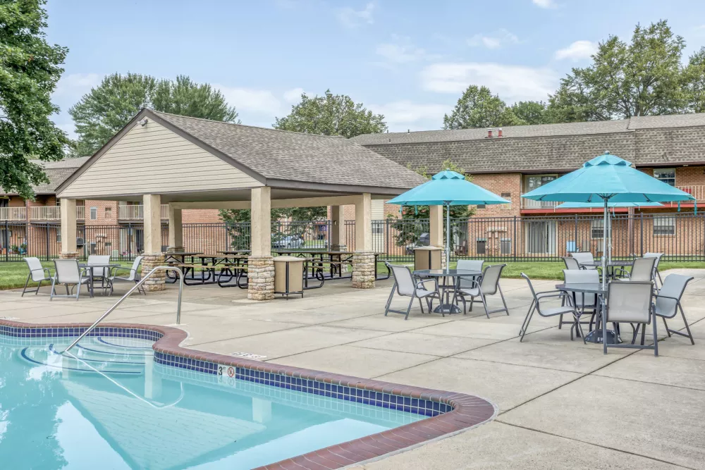 Apartment complex pool area with patio tables, two blue umbrellas, and a large covered pavilion.