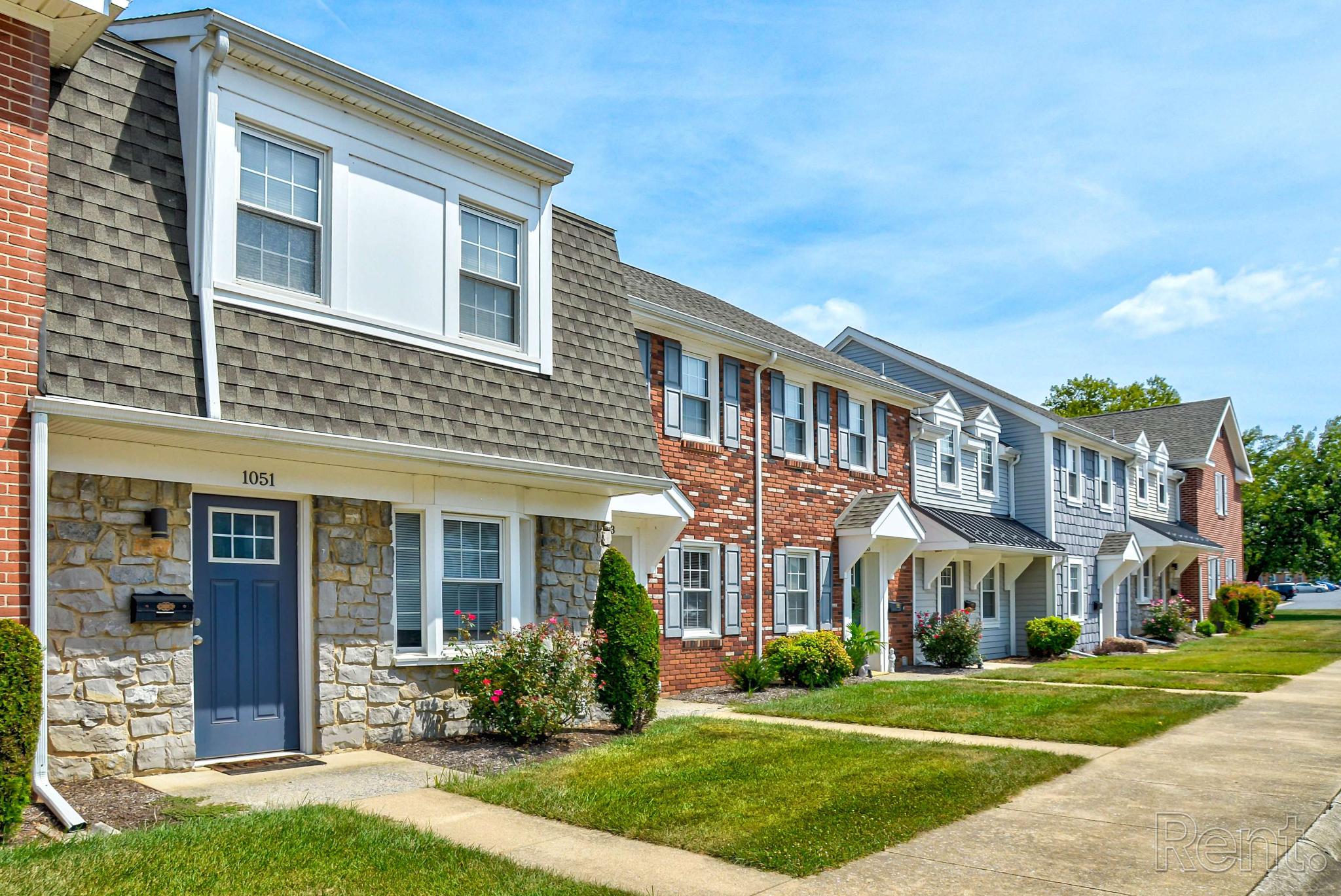 Row of brick and siding townhomes with gabled roofs and private entrances at Village of Olde Hickory Apartments, set along a clean sidewalk with manicured lawns and blooming shrubs.