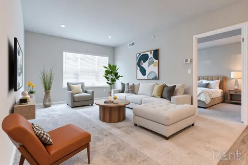 Modern luxury apartment living room with light gray walls, plush seating, a round wood coffee table, an orange leather accent chair, and a clear view into the adjacent bedroom.