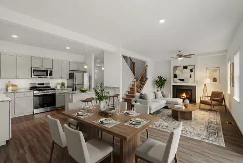 A wide-angle view of a modern open-concept living area featuring a kitchen with light grey cabinetry, granite countertops, and stainless steel appliances. The space flows into a dining area and a living room with a fireplace and large windows.