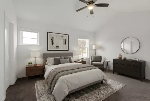 A spacious primary bedroom with neutral carpeting, white walls, and a large window. The room is styled with a tray ceiling and a modern ceiling fan.